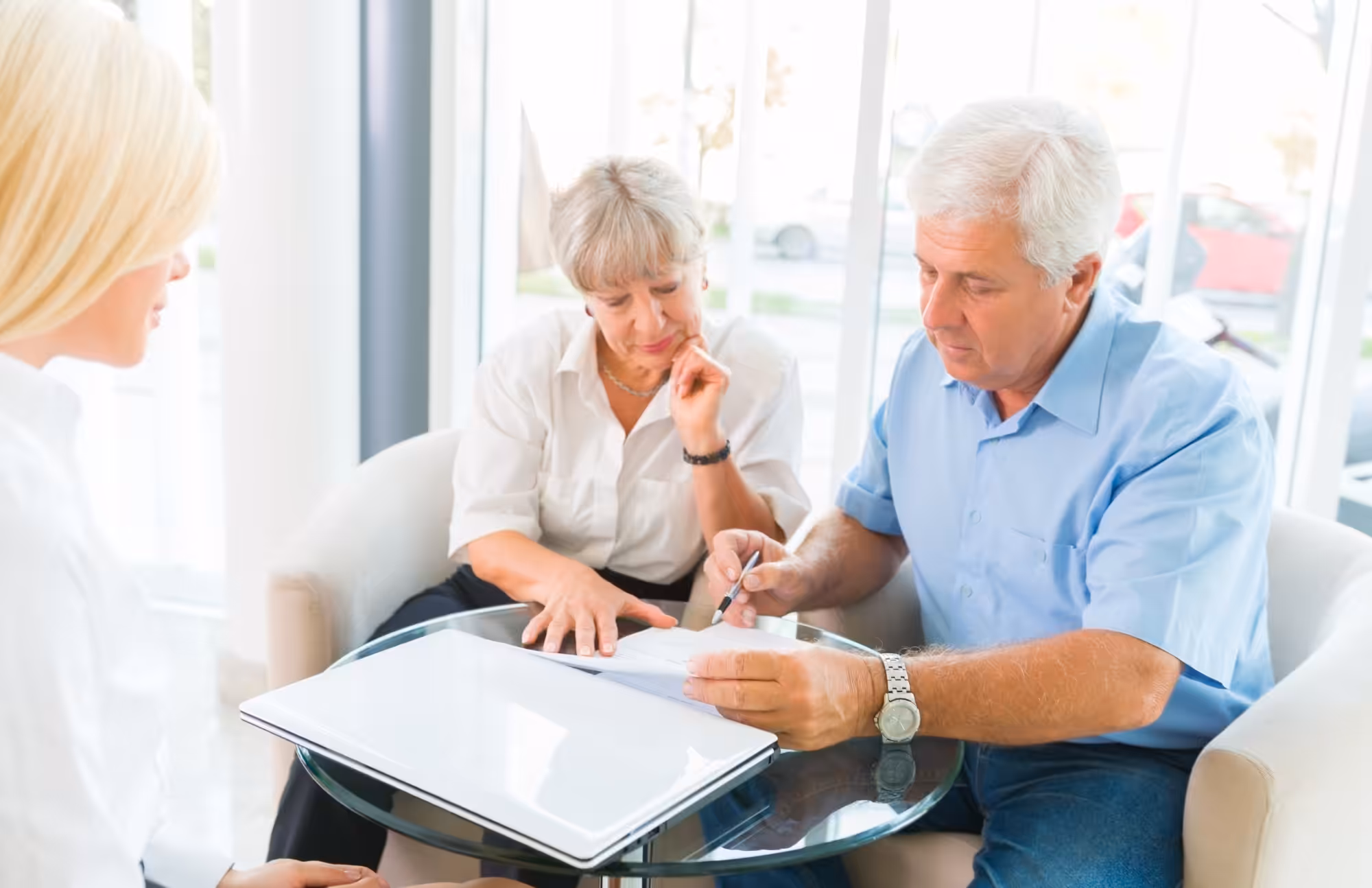An older couple sits at a glass table with a younger woman. The man signs a document while the woman looks on thoughtfully. Papers and a laptop are on the table, and large windows let in natural light.