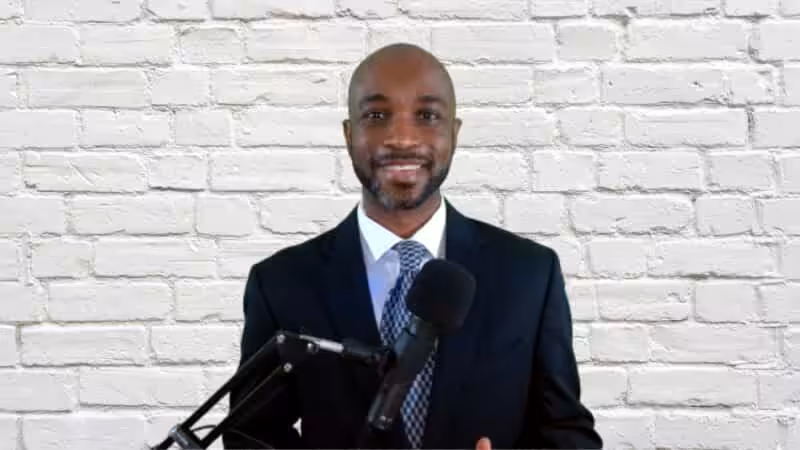 A man in a dark suit and tie stands in front of a white brick wall, smiling at the camera. A microphone on a stand is positioned in front of him, suggesting he is speaking or recording.