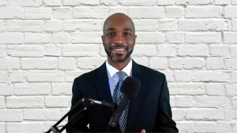 A man in a dark suit and tie stands in front of a white brick wall, smiling at the camera. A microphone on a stand is positioned in front of him, suggesting he is speaking or recording.
