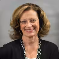 Smiling woman with shoulder-length wavy hair, wearing glasses, hoop earrings, a silver circular chain necklace, and a dark top, posing in front of a plain gray background.