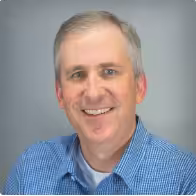 A smiling man with short gray hair wearing a blue checkered shirt and a white t-shirt underneath, posing against a plain gray background.