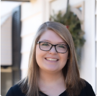 A young woman with straight, light brown hair and glasses smiles at the camera. She is wearing a black top and standing outdoors in front of a building with a green wreath on the door.