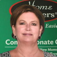 A woman with short brown hair and hoop earrings smiles at the camera. She stands in front of a green background with partially visible white text and a black oval logo.
