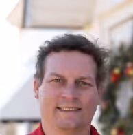 A man with short, wavy brown hair is smiling outdoors. He is wearing a red shirt, and there is a decorated wreath with red and gold ornaments on the wall behind him. The background is softly blurred.