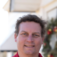 A man with short, wavy brown hair is smiling outdoors. He is wearing a red shirt, and there is a decorated wreath with red and gold ornaments on the wall behind him. The background is softly blurred.