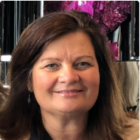 A woman with long brown hair smiles at the camera. She is indoors, and the background features pink flowers and reflective surfaces.