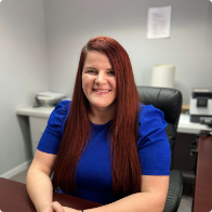 A woman with long straight red hair, wearing a blue top, sits and smiles at a desk in an office setting.