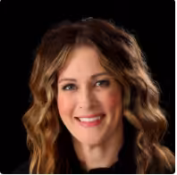 A woman with long, wavy brown hair and light highlights smiles at the camera against a black background. She is wearing a dark top.