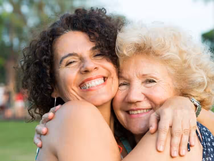 Two women smiling and hugging each other outdoors, showing happiness and affection. One has curly dark hair, the other has short blonde hair. The background is blurred greenery.