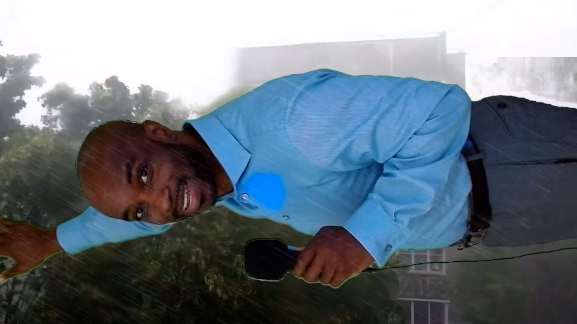 A man in a blue shirt, holding a microphone, leans sideways as if bracing against strong wind and rain outdoors, with trees and a blurry building in the background.