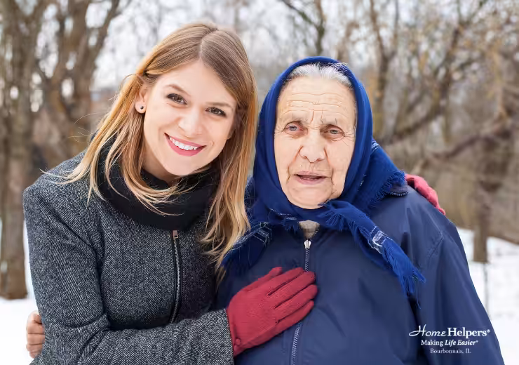A young woman smiles and stands close to an elderly woman in a blue coat and headscarf. They are outdoors in a snowy setting. The younger woman wears a gray coat and red gloves. Home Helpers logo is visible in the corner.
