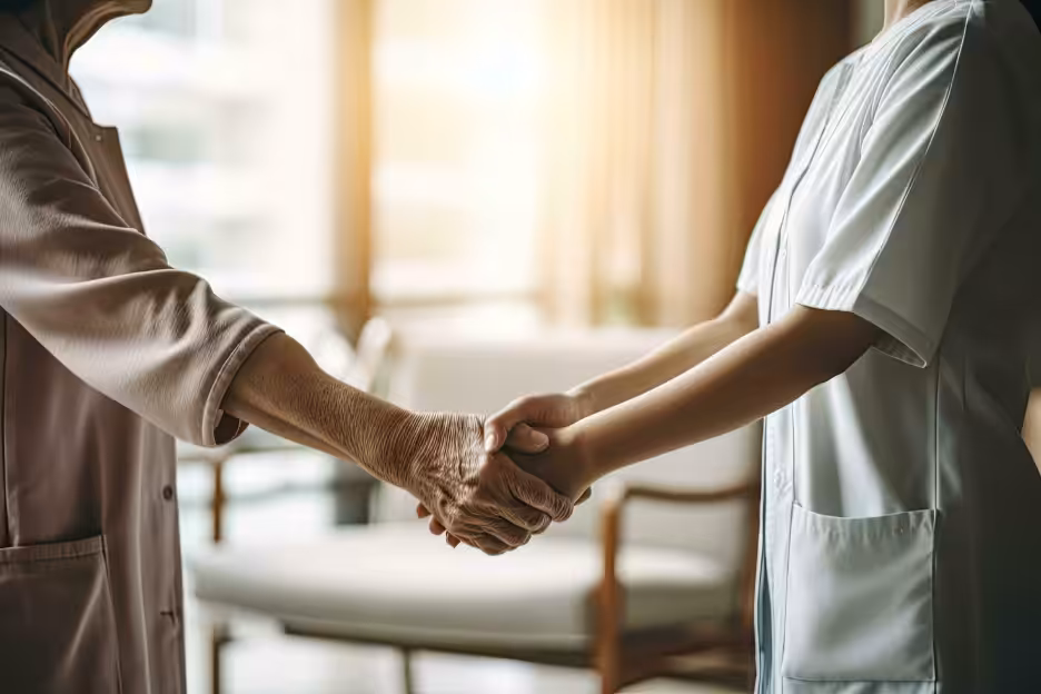 An elderly person and a healthcare worker stand indoors, holding hands in a supportive gesture, with soft natural light filling the background.