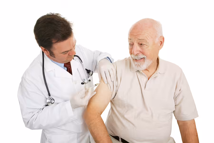 A doctor wearing a white coat and gloves gives a vaccine injection in the upper arm of an older man with a beard, who is sitting and wearing a beige polo shirt, against a plain white background.