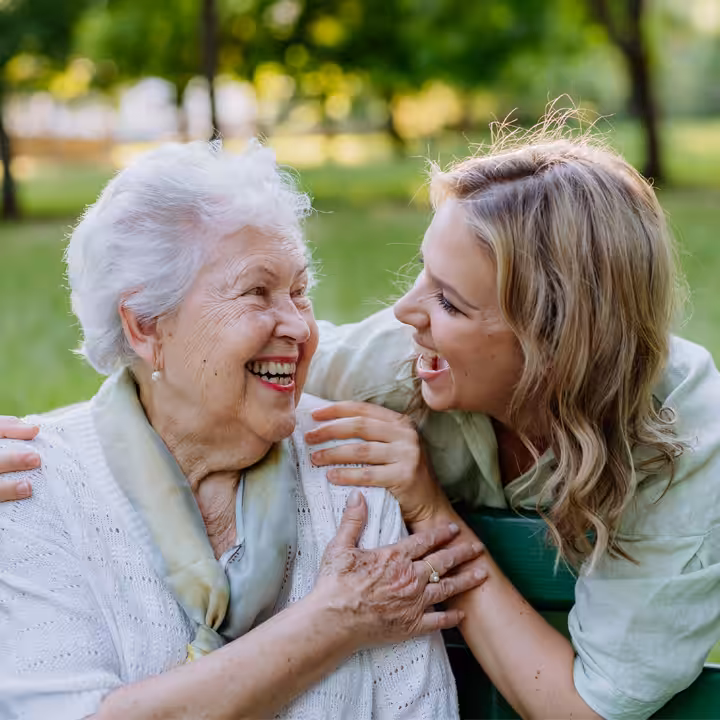 An elderly woman and a younger woman sit outdoors on a bench, smiling and laughing together. The background is green and leafy, suggesting a park or garden setting. They appear happy and enjoy each others company.
