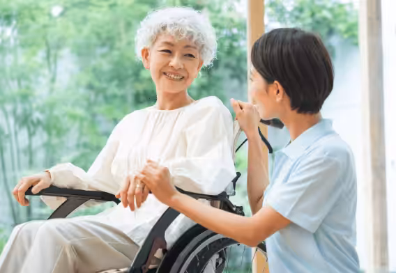 An older woman with curly gray hair sits in a wheelchair, smiling warmly at a younger woman in a light blue uniform, who is holding her hand and talking to her indoors by a window with greenery outside.