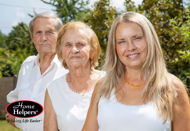 Three people stand outdoors: an elderly man, an elderly woman, and a middle-aged woman smiling at the camera. The “Home Helpers” logo is in the lower left corner, along with the tagline Making Life Easier.