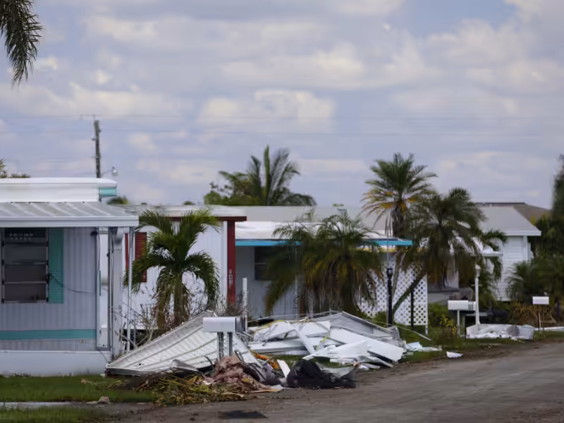 Damaged mobile homes with debris and broken panels scattered in front, palm trees and other homes visible in the background under a partly cloudy sky.
