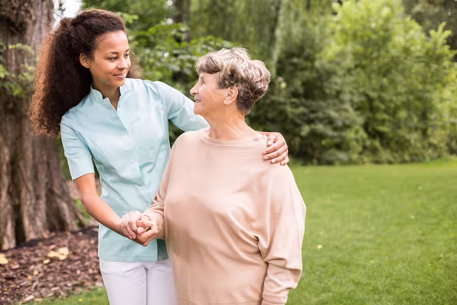 A young caregiver smiles and supports an older woman as they walk together outdoors in a green, leafy park. The caregiver has her arm around the woman, and both appear happy and engaged.