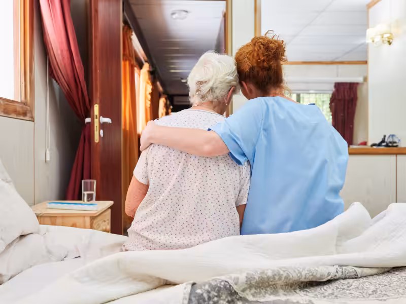 A caregiver in blue scrubs sits on a bed beside an elderly woman, gently putting an arm around her shoulders in a supportive gesture. They are in a cozy, well-lit room with red curtains and a bedside table.