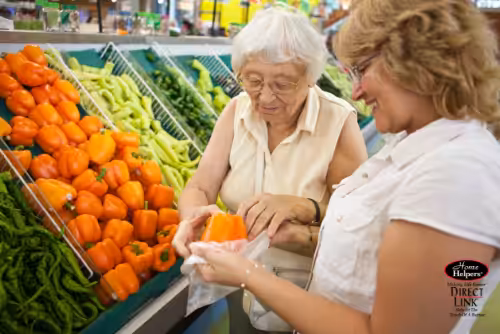 Two women stand at a grocery store produce section, selecting orange bell peppers together. One woman holds a pepper while the other helps put it in a plastic bag. Green and orange peppers are displayed around them.