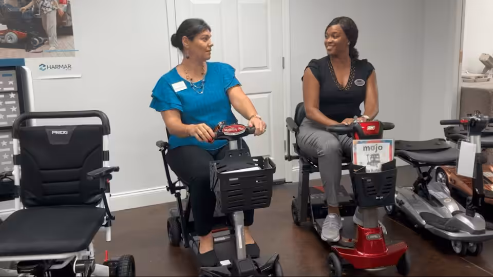 Two women sit indoors on mobility scooters, facing each other and smiling. They appear to be in a showroom with other scooters displayed around them and posters on the wall in the background.