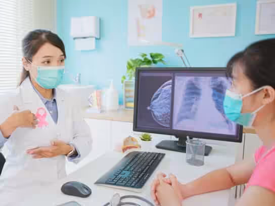 A doctor wearing a mask and a pink ribbon discusses breast health with a masked patient. X-ray and mammogram images are displayed on a computer screen in the background.