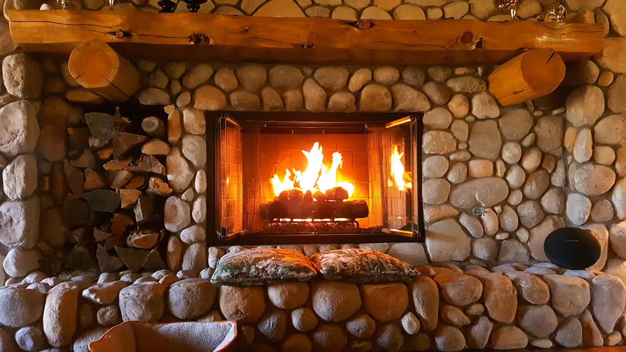 A cozy stone fireplace with a blazing fire, stacked logs on the left, two patterned cushions on the stone hearth, and a rustic wooden mantel above.