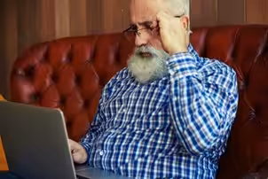 An older man with a white beard and glasses, wearing a blue plaid shirt, sits on a brown leather couch looking intently at a laptop screen.