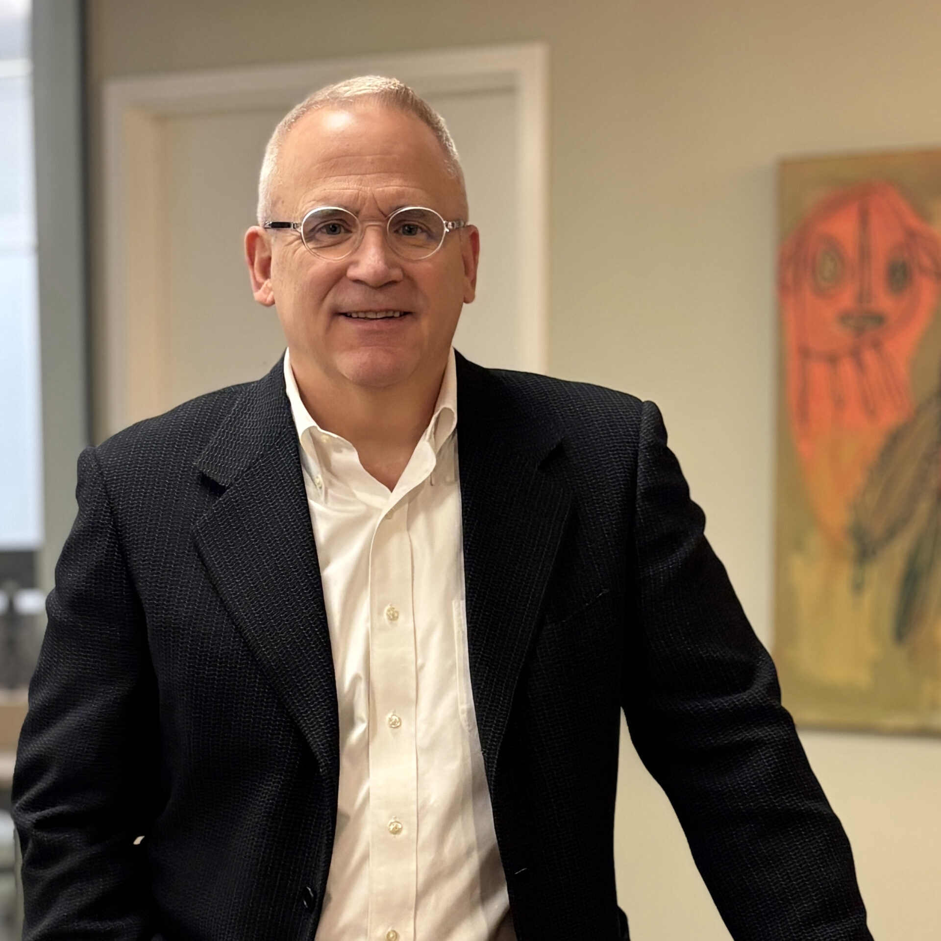 A man with short gray hair and glasses, wearing a dark blazer over a white shirt, stands smiling in an office setting. A colorful abstract painting is visible on the wall behind him.