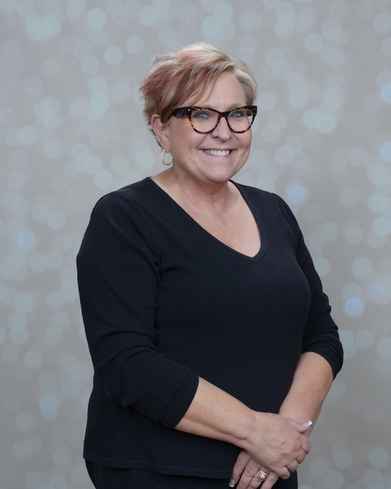 A woman with short, light hair and glasses is smiling, wearing a black V-neck top and gold hoop earrings, standing in front of a light, blurred background with circular bokeh lights.