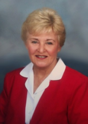 A smiling older woman with short blonde hair is wearing a red blazer over a white collared shirt, posing in front of a soft, cloudy blue-gray background.