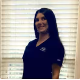 A woman with long dark hair wearing a black shirt stands indoors in front of white window blinds, smiling at the camera.
