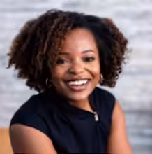 A woman with curly hair, wearing a black sleeveless top, smiles warmly while sitting in front of a light gray, textured background.