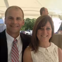 A man in a suit and striped tie sits next to a woman in a white sleeveless dress. They are smiling under a canopy at an outdoor event, with another man and greenery visible in the background.