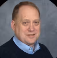 A middle-aged man with short light hair, wearing a navy sweater over a light blue collared shirt, smiles at the camera against a gray background.