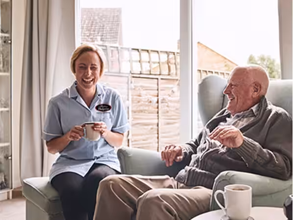 A smiling caregiver sits beside an elderly man in a cozy living room, both holding mugs and enjoying a conversation near a large window with sunlight streaming in.