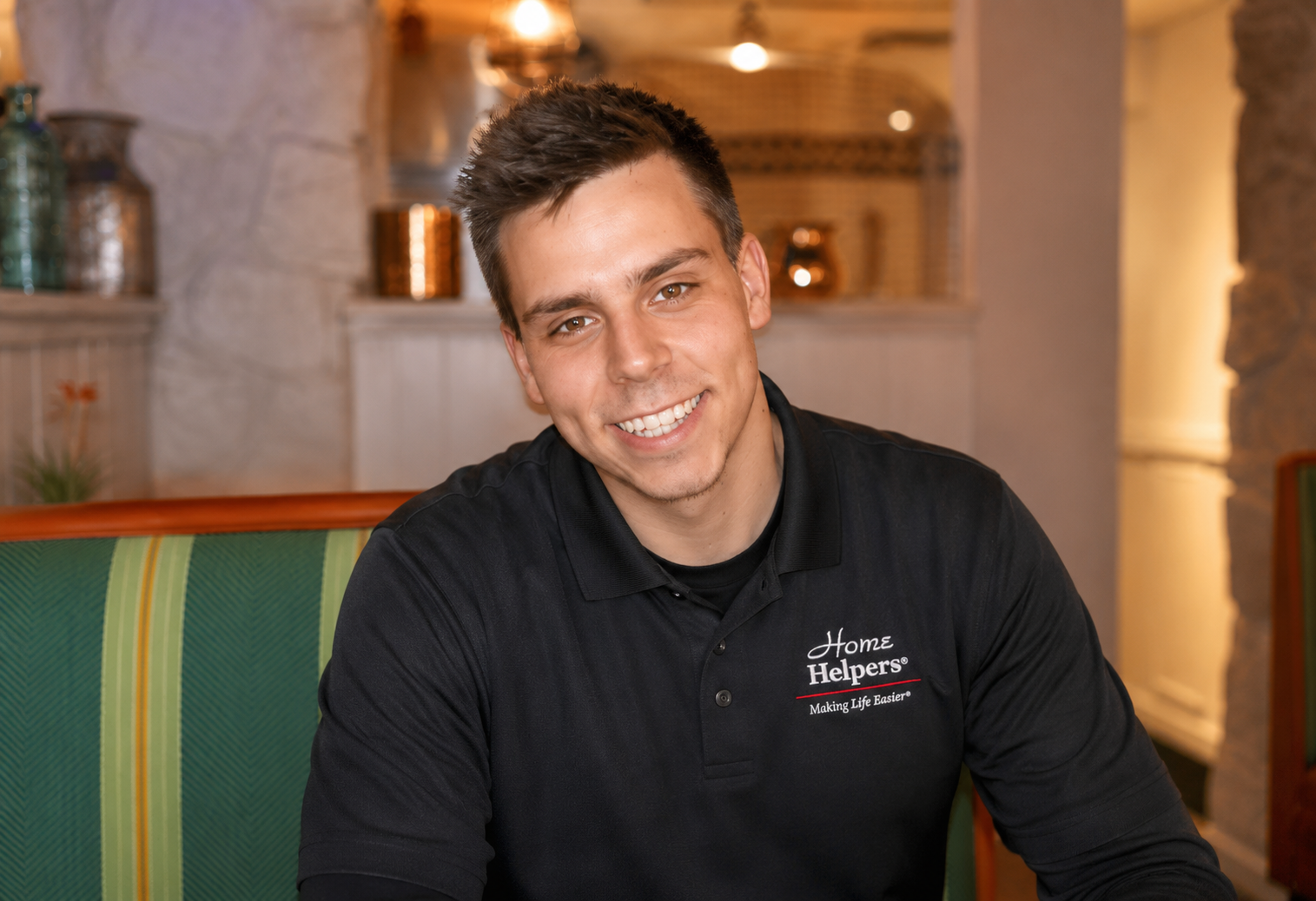 A young man wearing a black “Home Helpers” polo shirt sits on a green striped bench, smiling at the camera in a warmly lit room with stone and wooden decor in the background.