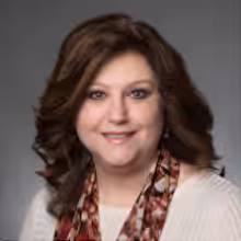 A woman with wavy brown hair smiles at the camera. She is wearing a cream-colored top and a patterned scarf, in front of a plain gray background.