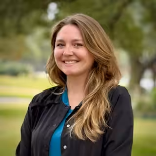 A woman with long, light brown hair wearing a black jacket over a teal top smiles outdoors with trees and greenery blurred in the background.