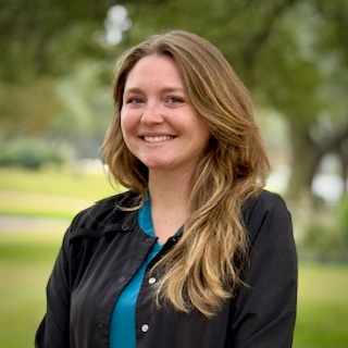 A woman with long, light brown hair wearing a black jacket over a teal top smiles outdoors with trees and greenery blurred in the background.