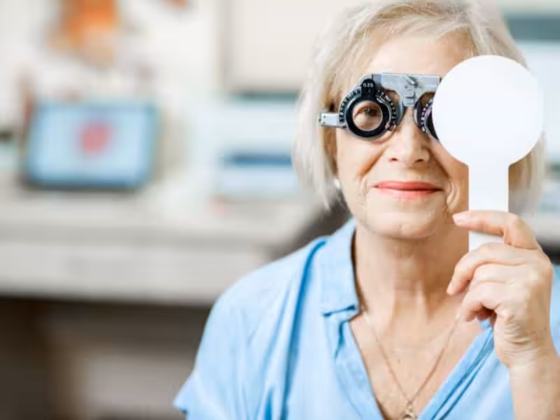 An older woman wearing a blue shirt sits in an eye clinic. She is using a phoropter for an eye exam and holds a white occluder over one eye. The background is blurred with medical equipment visible.