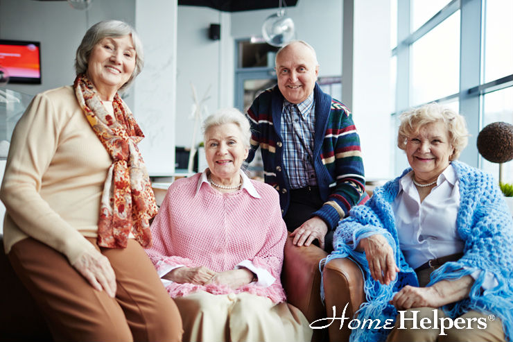 Four smiling older adults, three women and one man, sit together indoors. They are dressed in cozy clothing and appear happy and relaxed. The Home Helpers logo is visible in the bottom right corner.