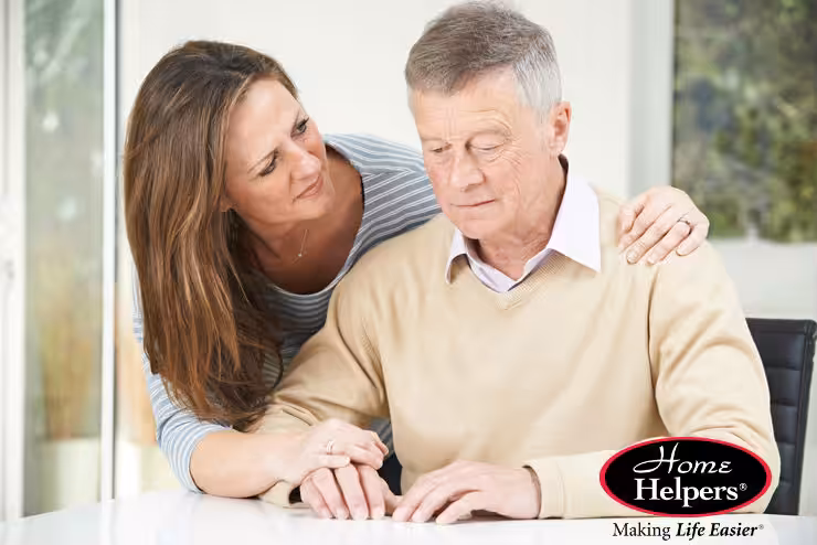 A woman gently supports an elderly man who is sitting at a table, looking down thoughtfully. The image includes the Home Helpers logo and the slogan “Making Life Easier” in the bottom right corner.