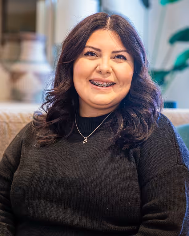 A woman with long dark hair and braces smiles warmly while sitting indoors, wearing a black sweater and a necklace. The background is softly blurred with some plants visible.