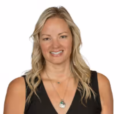A woman with long, wavy blonde hair wearing a sleeveless black top and a necklace with a circular pendant, smiling against a white background.
