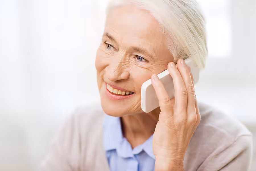 Smiling older woman with gray hair holds a smartphone to her ear, appearing happy while talking on the phone. She wears a light blue blouse and light-colored sweater in a bright, softly-lit setting.