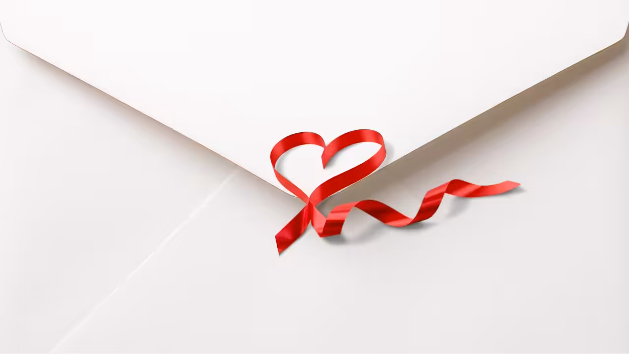 A white envelope is sealed with a red ribbon shaped into a heart and trailing off in a curl against a white background.