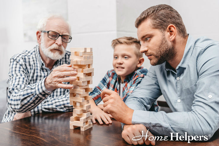 Three generations of men—a grandfather, father, and young boy—are playing a game of Jenga at a table, focused on carefully removing blocks. Home Helpers logo appears in the bottom right corner.