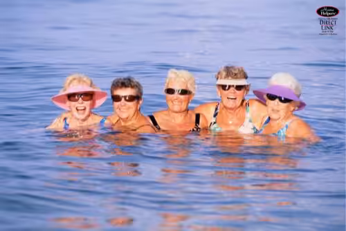 Five older women in swimsuits and sunglasses, smiling and standing close together in calm blue water, enjoying a sunny day.