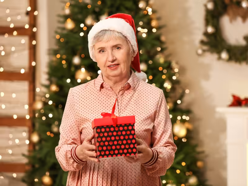An older woman wearing a Santa hat and a pink polka-dot shirt smiles while holding a red and black gift box in front of a decorated Christmas tree with lights in the background.